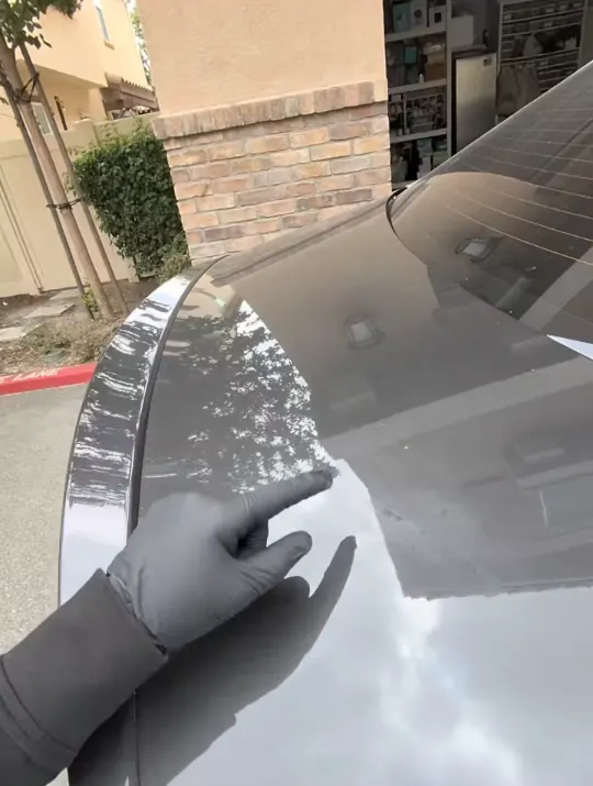 Gloved hand pointing to a scratch on the trunk lid of a dark sedan